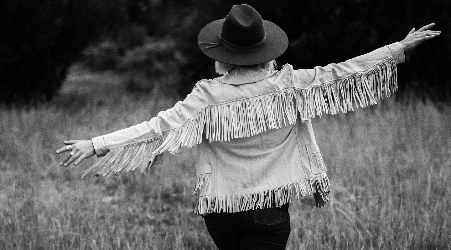 Person wearing a fringed jacket and hat with arms outstretched in a field